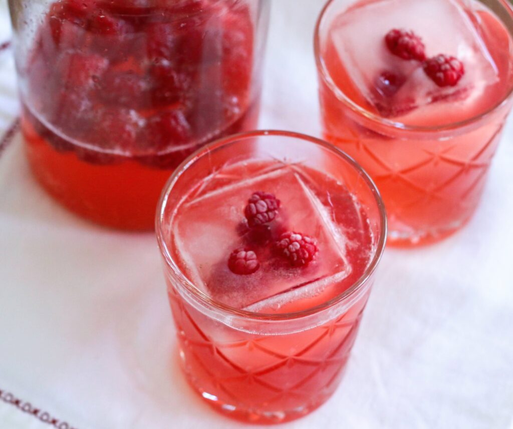 A glass of red liquid with ice cubes and raspberries.